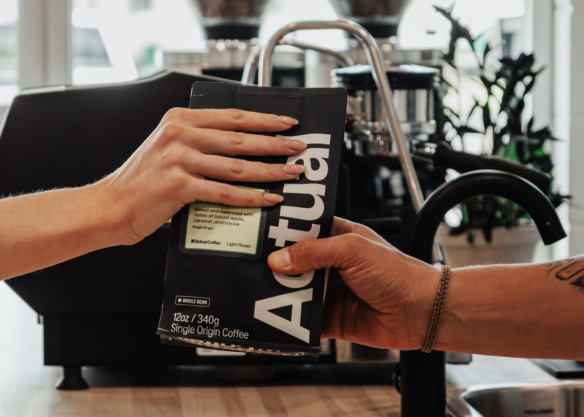 Person holding a bag of Actual Coffee in a kitchen setting with coffee grinders in the background.