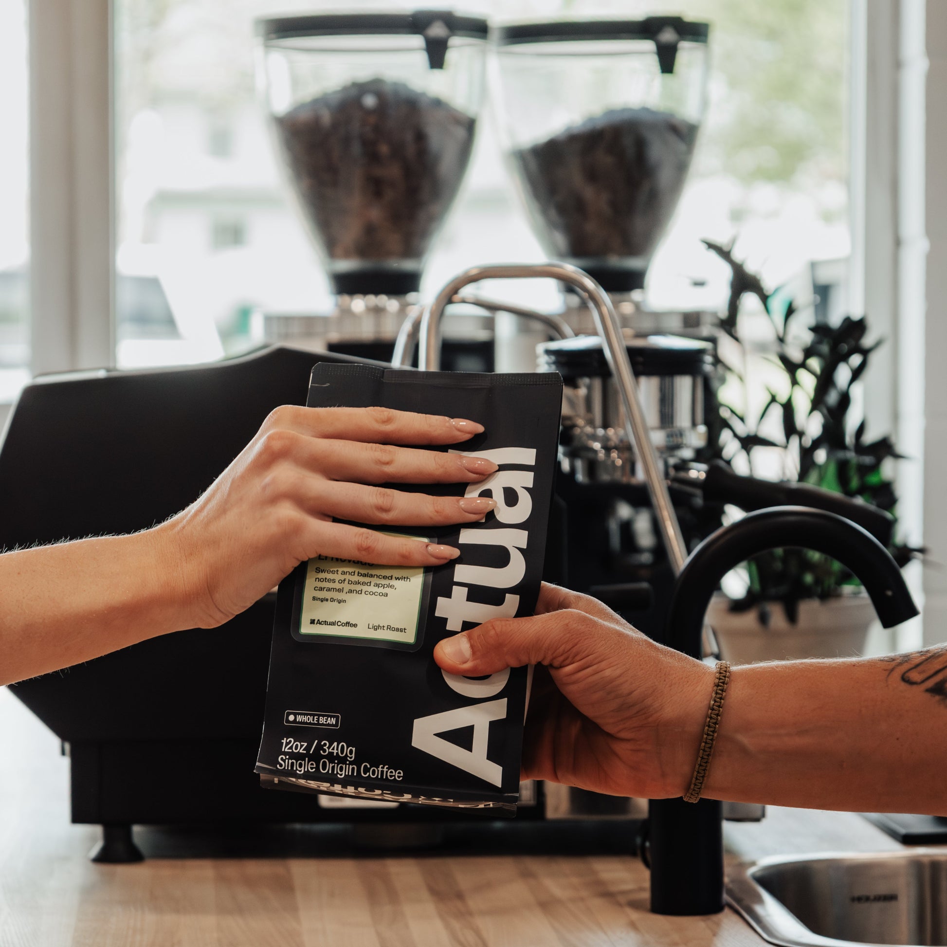 Person holding a bag of Actual Coffee in a kitchen setting with coffee grinders in the background.
