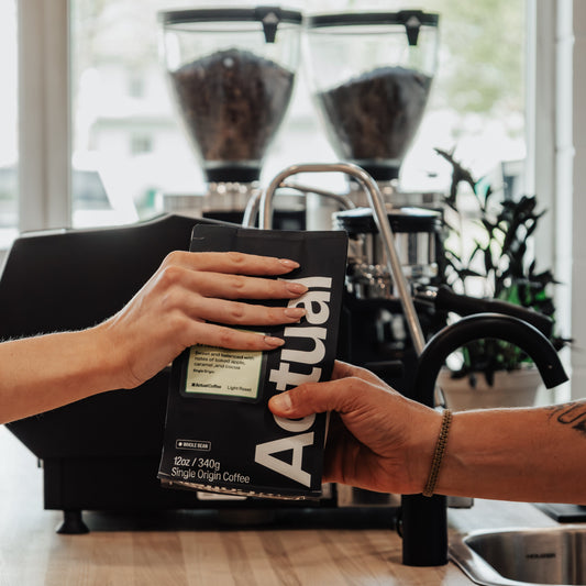 Person holding a bag of Actual Coffee in a kitchen setting with coffee grinders in the background.